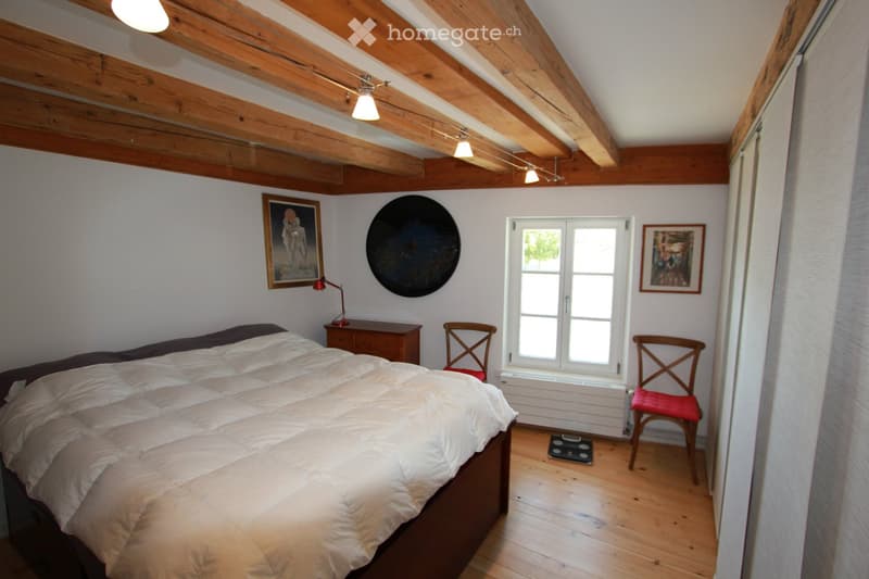 Cozy bedroom with wooden beams, a large bed with white bedding, a nightstand with a lamp, two wooden chairs, and artwork on the walls.