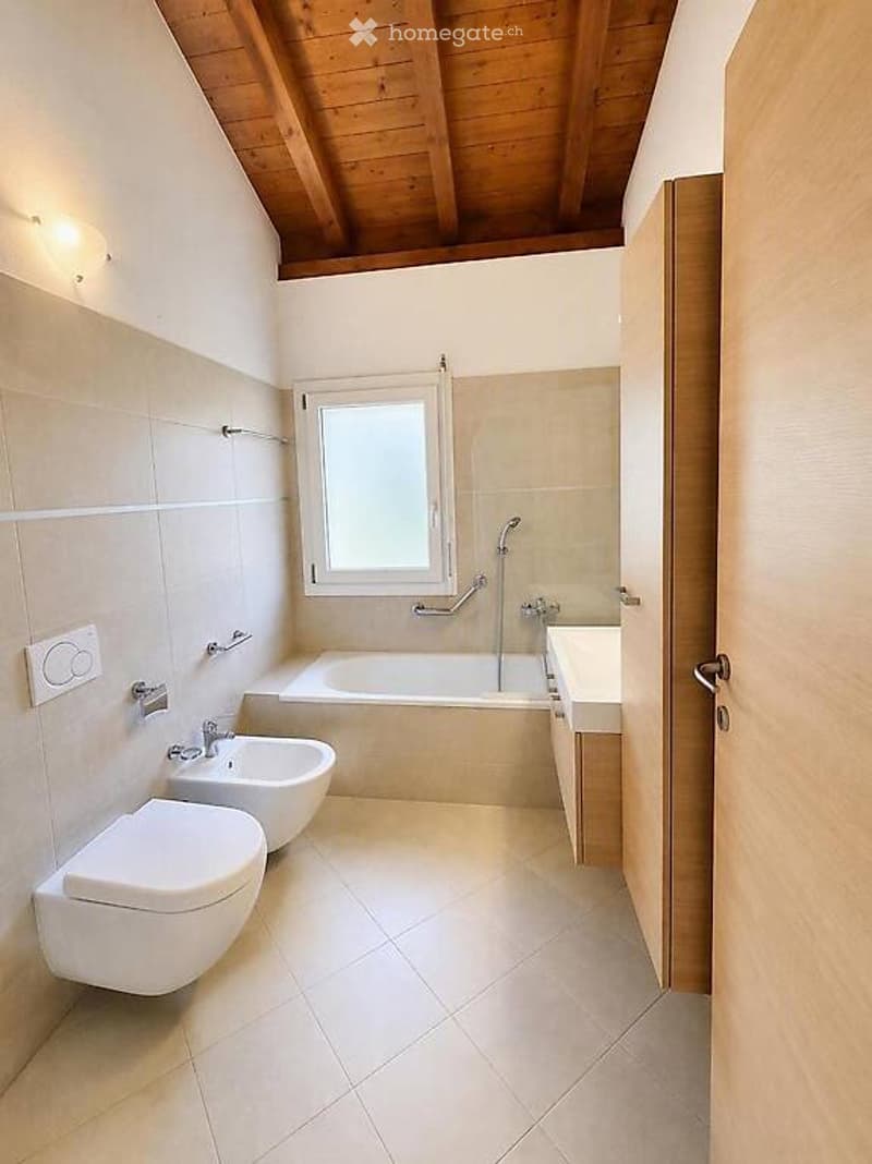 Modern bathroom with white tiles, a bathtub, toilet, bidet, and sink. Features a wooden ceiling and a small window above the bathtub.