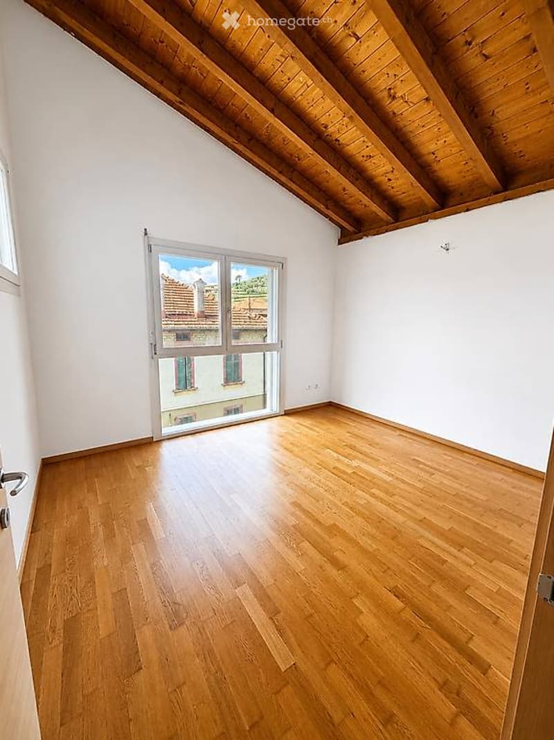 Empty room with wooden flooring, white walls, and large windows offering a view of the outside. The ceiling has exposed wooden beams.