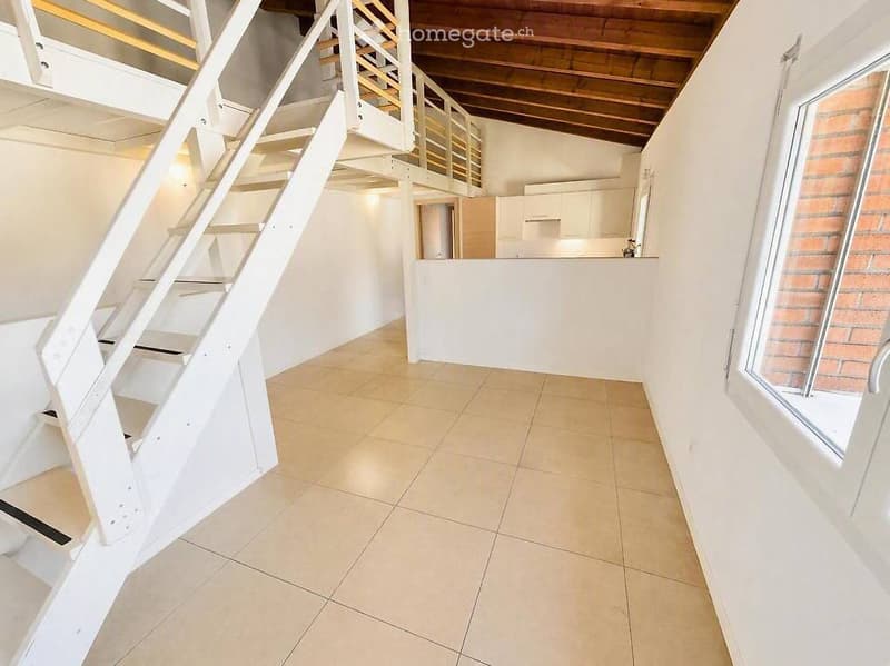 Open-plan space with a staircase leading to an upper level, tiled floor, white walls, and a small kitchen area with cabinets and a countertop.