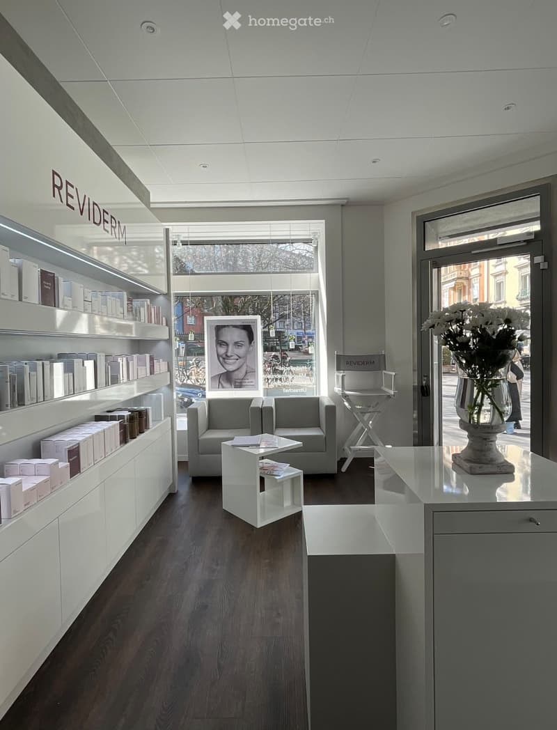 Modern, minimalist interior with a grey sofa, white coffee table, shelves with product boxes, and a vase with flowers near the entrance.