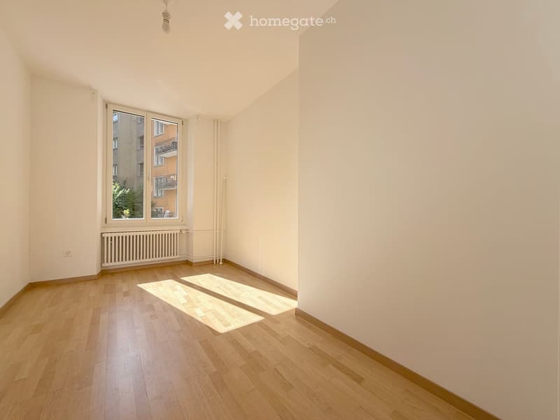 Empty room with wooden flooring, white walls, and large windows showing a view of the outside buildings.
