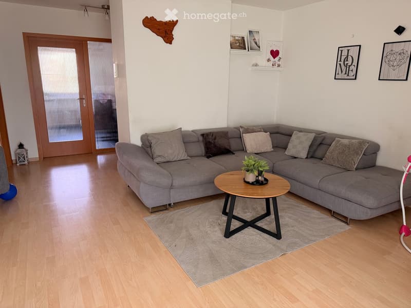 Modern living room with a grey sectional sofa, wooden coffee table, potted plants, and a grey rug on a light wooden floor.