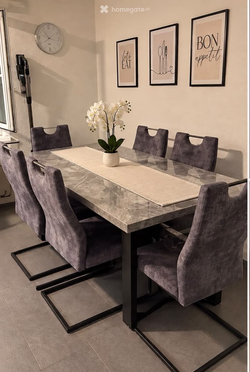 Dining room with a marble table, six grey upholstered chairs, a white flower vase, a wall clock, and three picture frames on the wall.