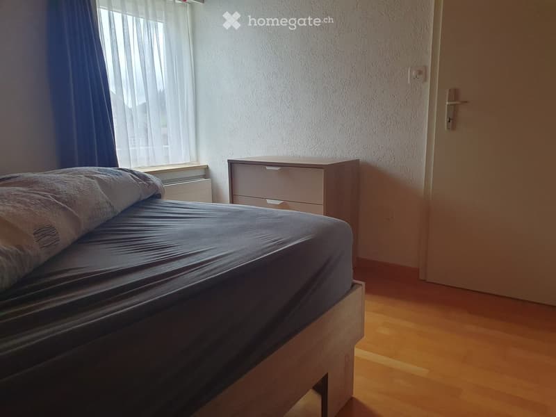 A small bedroom with a single bed, a nightstand, and a drawer unit. The room has wooden flooring and a window with sheer curtains.