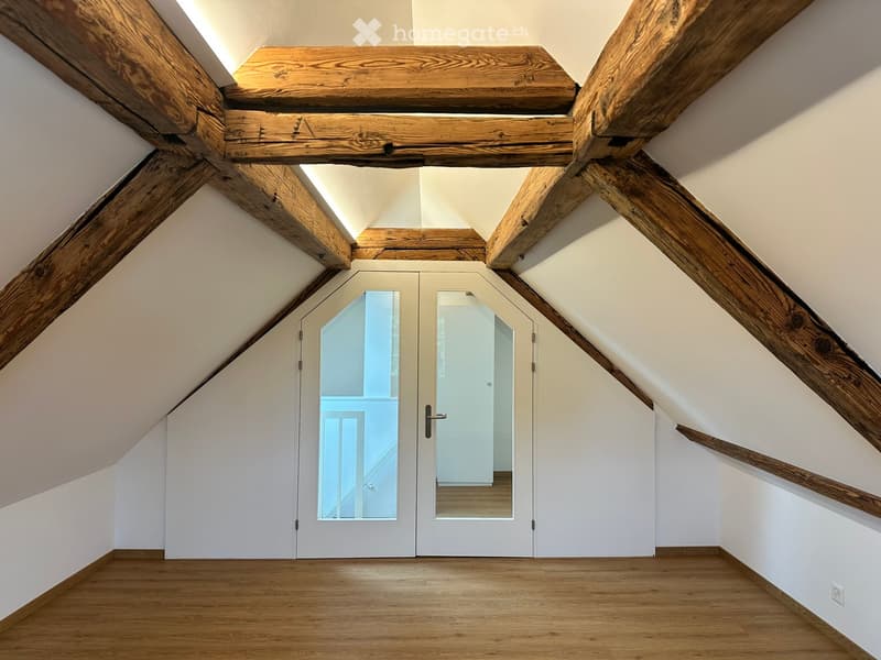 An attic space with exposed wooden beams, white walls, and a wooden floor. A set of white double doors with glass panels leads to another room.
