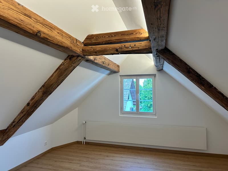 An attic room with wooden beams, wooden flooring, white walls, and a window offering a view of the roof and greenery outside.