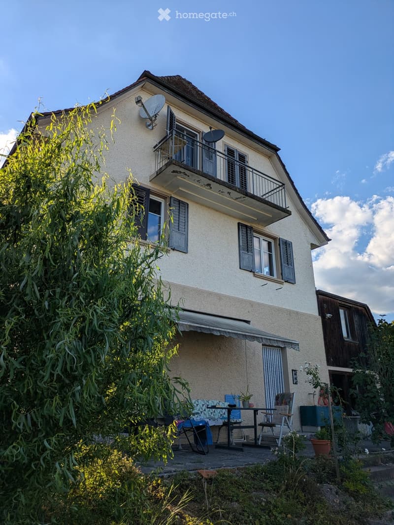 Two-story house with beige exterior, multiple windows, shutters, and a balcony on the upper floor. The lower level features a covered patio with outdoor furniture, including a table and chairs, surrounded by greenery and plants.