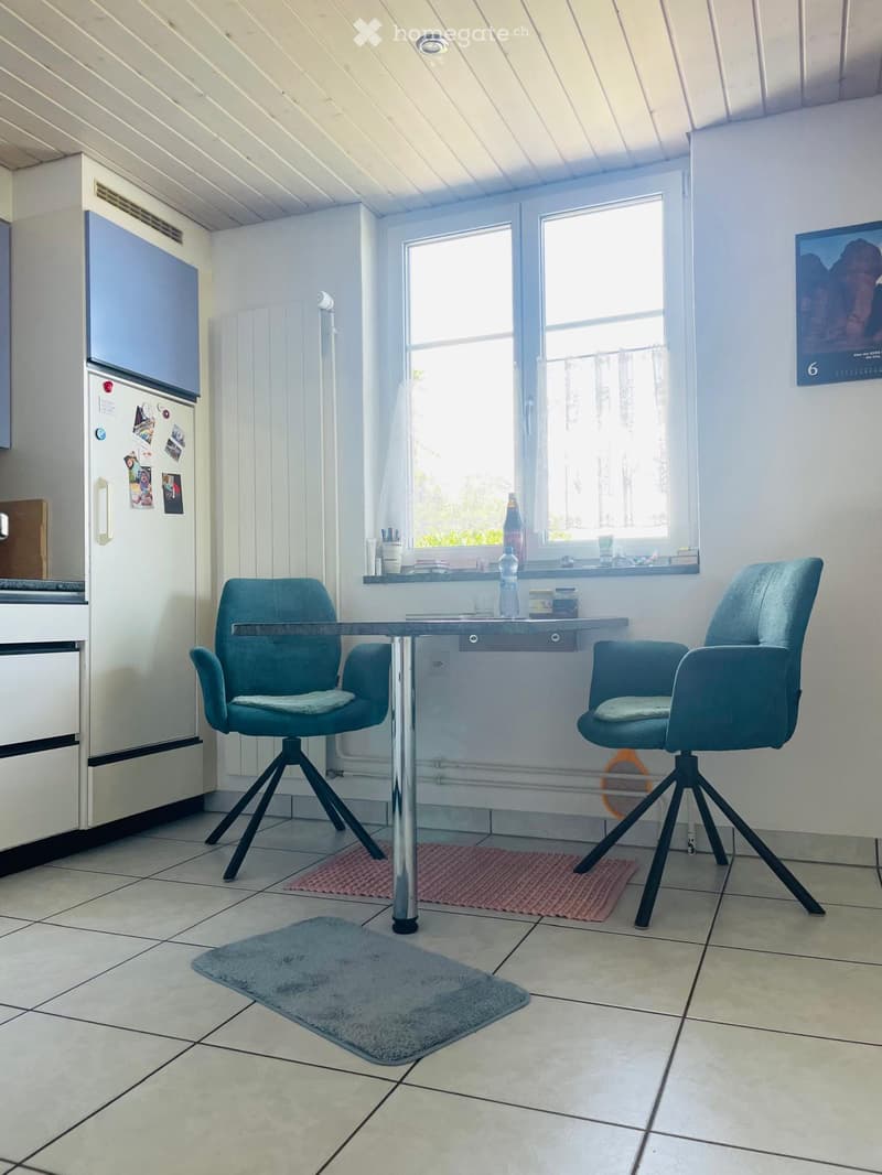 A small, modern kitchen area with a table, two chairs, a refrigerator, and a window with a view of greenery outside.