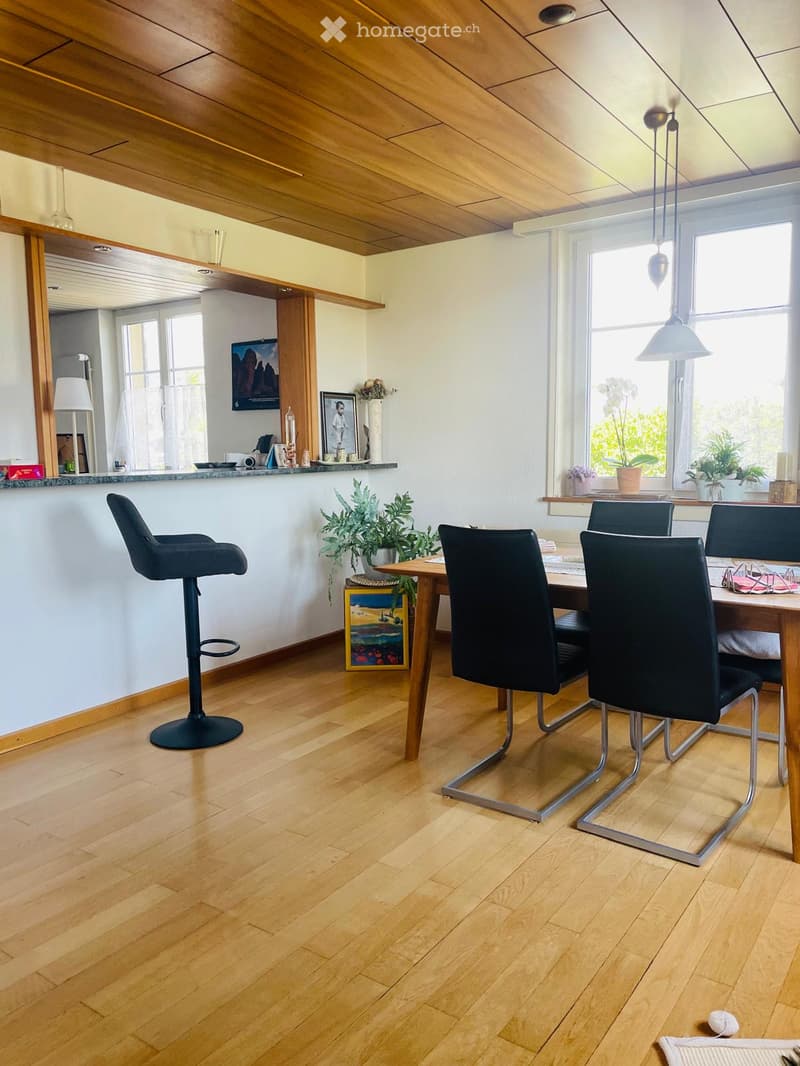Dining area with wooden floor, pendant lights, a wooden table with four black chairs, a black stool, and decorative plants and picture frames on the wall.