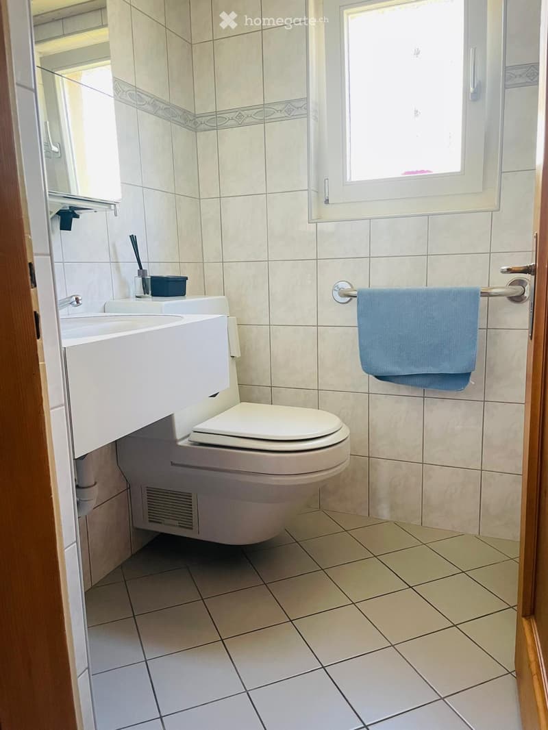 Modern bathroom with white tiles, a toilet, a sink with a faucet, a towel on a rack, and a toothbrush holder on the sink.