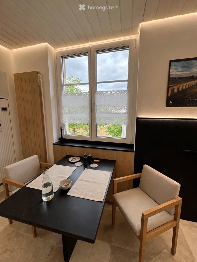 A modern, well-lit room with a black dining table, two chairs, a vase, and a bowl on the table. There are two windows with white curtains, a picture frame on the wall, and a sleek cabinet.