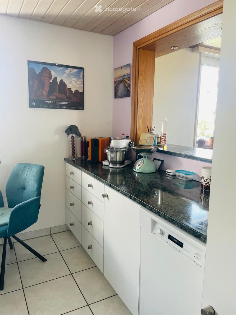 A modern kitchen with white cabinets, granite countertops, a dishwasher, a blender, a scale, a coffee machine, a toaster, and a chair. There are also various small appliances and decorative items on the counter.