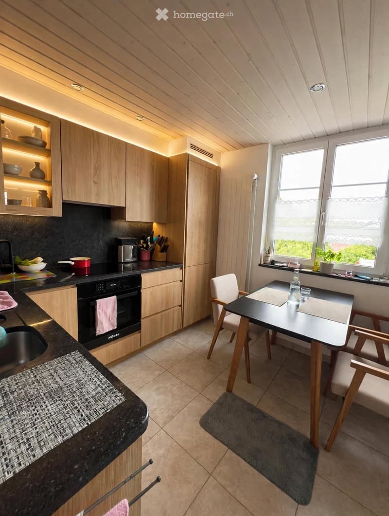 Modern kitchen with light wooden cabinets, black countertops, and built-in appliances. Includes a dining area with a table and chairs, a sink, and various kitchen items on the counters.