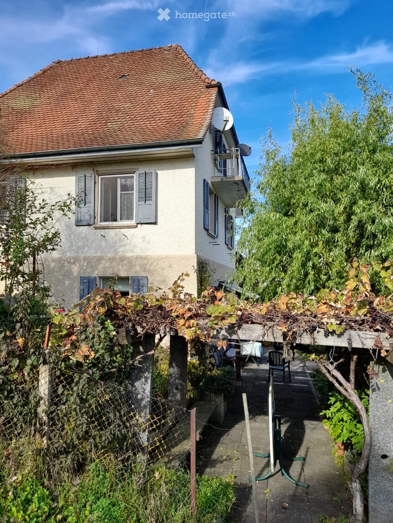 Two-story house with a red-tiled roof, white walls, and multiple windows. A balcony is visible on the upper floor, and the house is surrounded by lush greenery and grapevines.