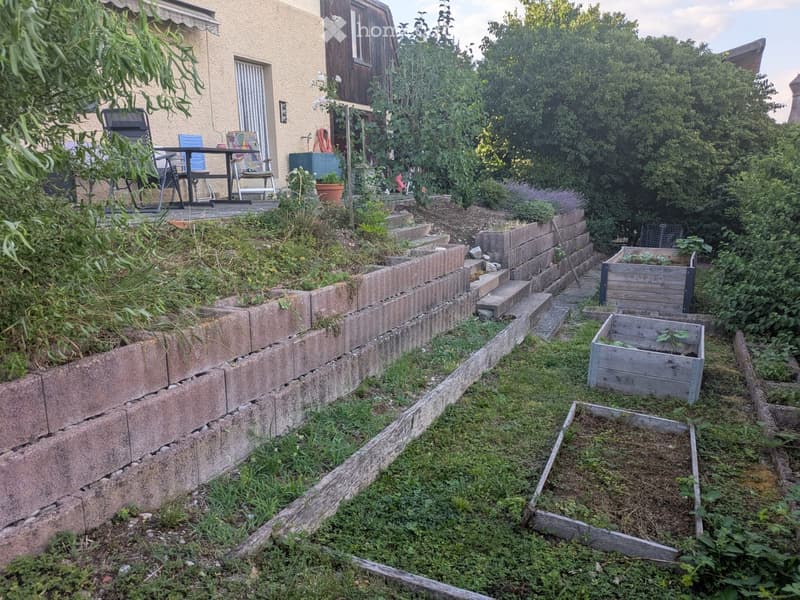 Garden with raised brick retaining wall, wooden planters, and outdoor seating area with table and chairs near the house.