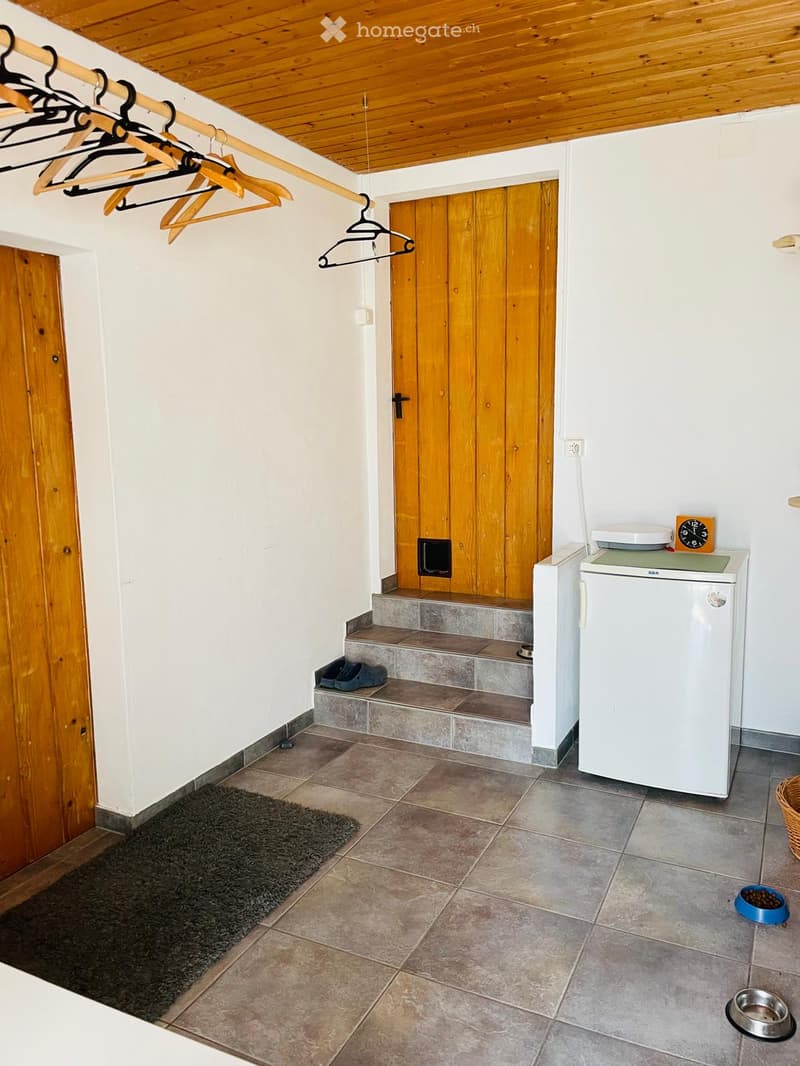 The image shows a small, well-lit hallway or entryway with tiled flooring, a wooden ceiling, and white walls. A small refrigerator is positioned against the wall, with a clock on top. A wooden coat rack with several hangers is mounted on the wall, and a s