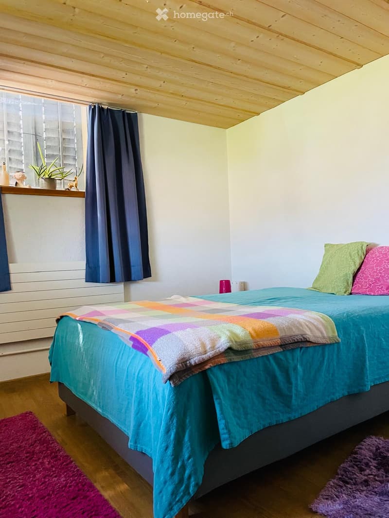 A cozy bedroom with a wooden bed, colorful blanket, pillows, and a window with dark curtains. A small plant sits on the windowsill, and colorful rugs are placed on the wooden floor.