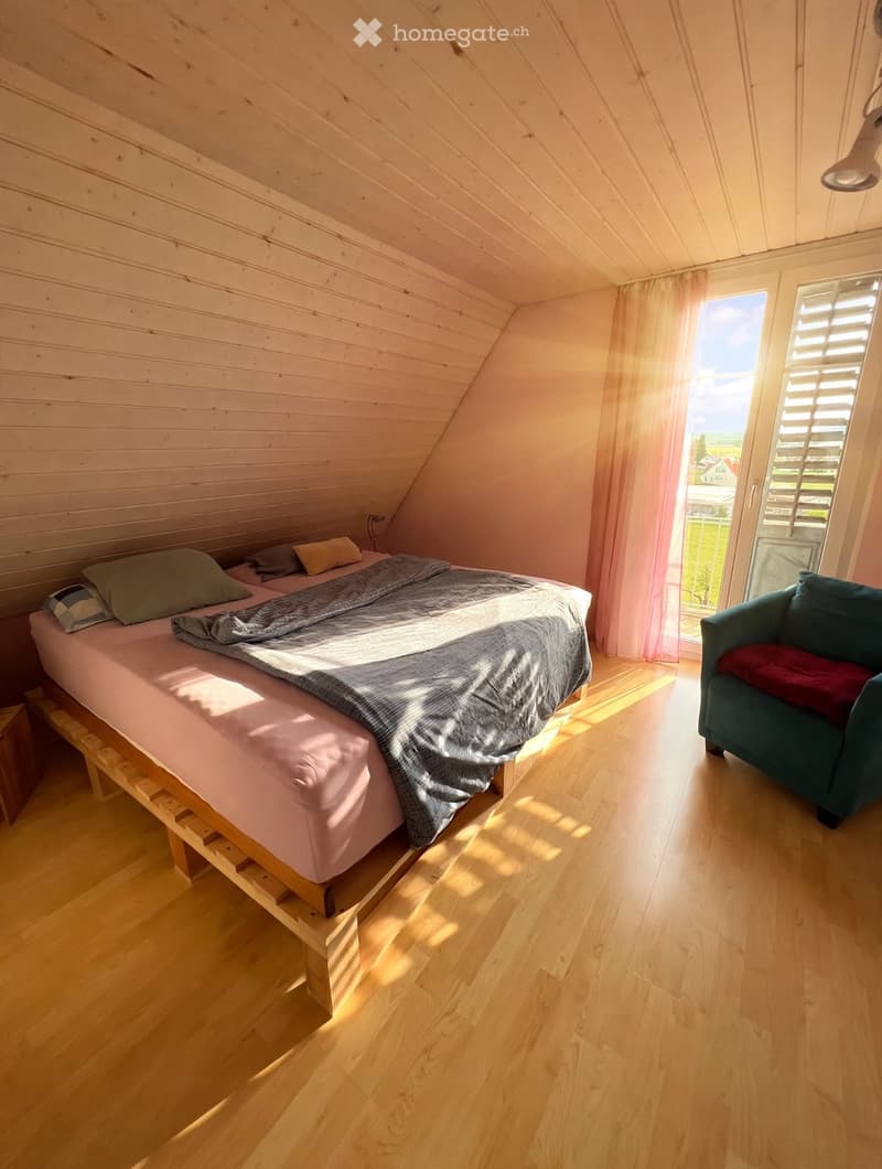 Cozy attic bedroom with wooden slanted ceilings, a single bed with pillows and a blanket, a small sofa, and large windows with sheer curtains letting in natural light.