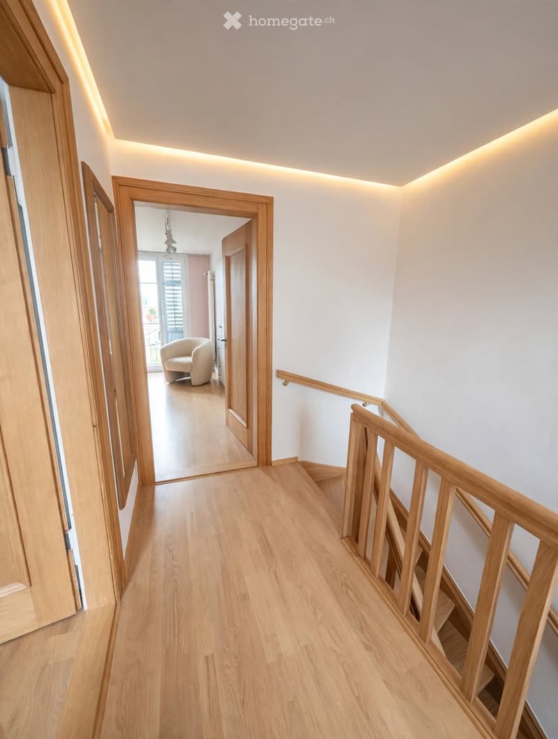 Spacious hallway with light wooden flooring, white walls, and recessed lighting. Features a staircase with wooden railings and open doorways leading to other rooms.