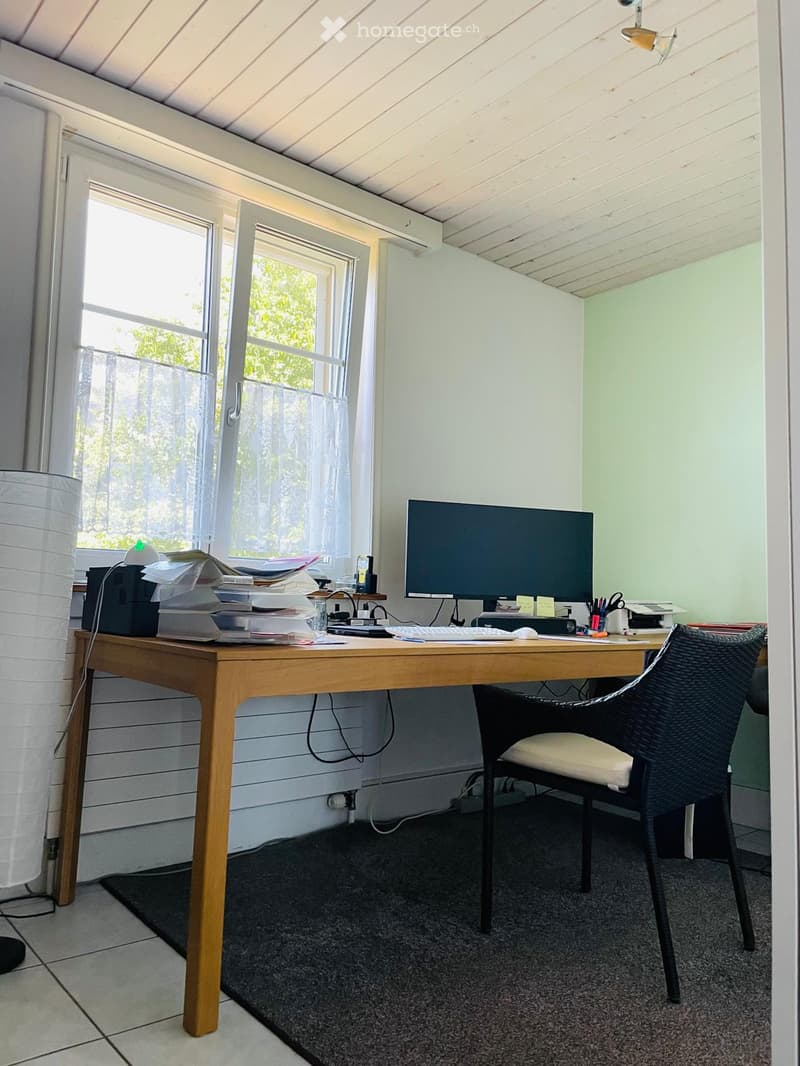 A well-lit study or home office with a wooden desk, computer setup, and a wicker chair. The room features large windows with curtains and a modern lamp.