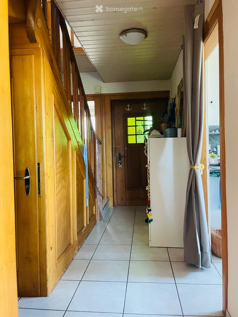 A well-lit hallway with tiled flooring, wooden railings, and a staircase leading upwards. The hallway features a closed wooden door at the end and a curtain on the right side.