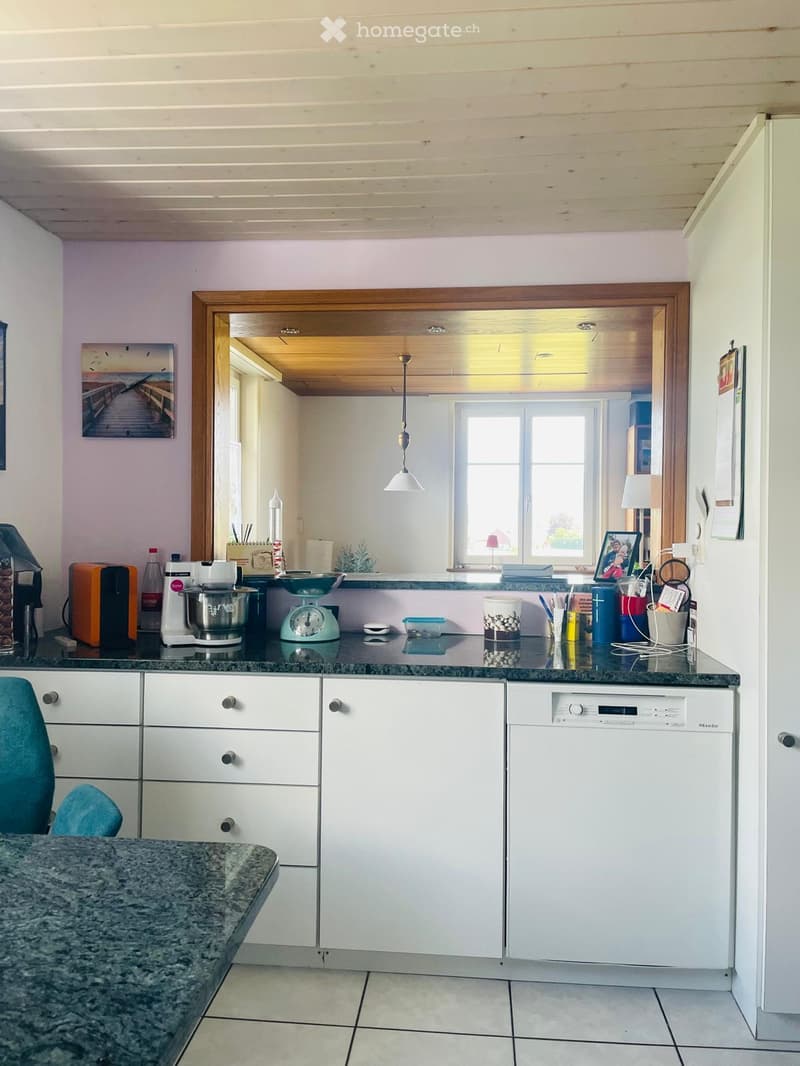 Modern kitchen with white cabinets, granite countertops, and various appliances including a dishwasher, coffee maker, mixer, and scale. A small dining area with a chair is visible in the foreground.