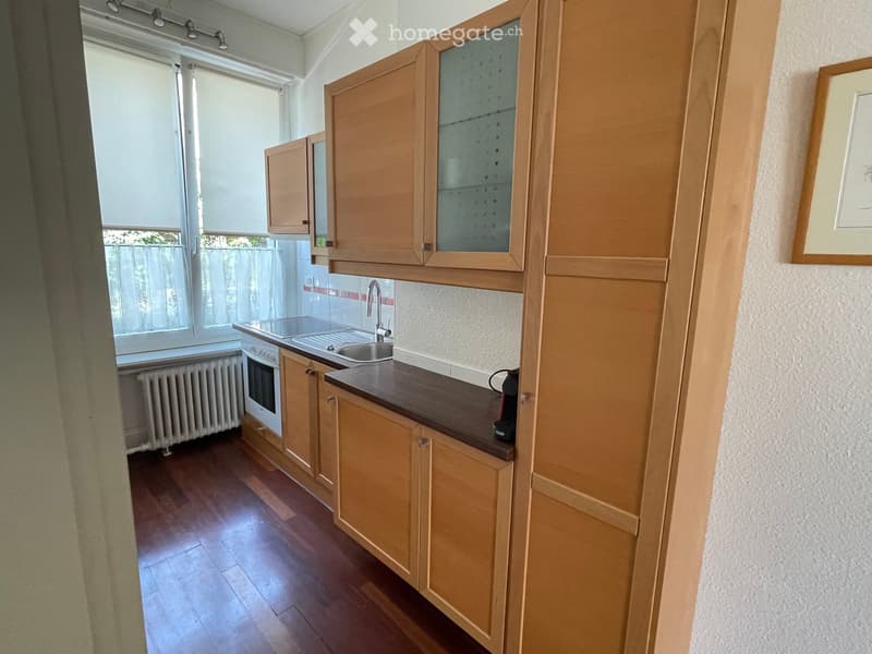 Modern kitchen with wooden cabinets, stainless steel sink, oven, and coffee maker on the counter. Natural light from large windows with blinds.