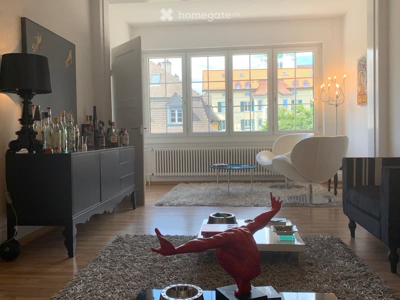 Modern living room with large windows, wooden floor, white shag rug, grey sofa, white armchair, glass coffee table, decorative lamp, and a cabinet with bottles.
