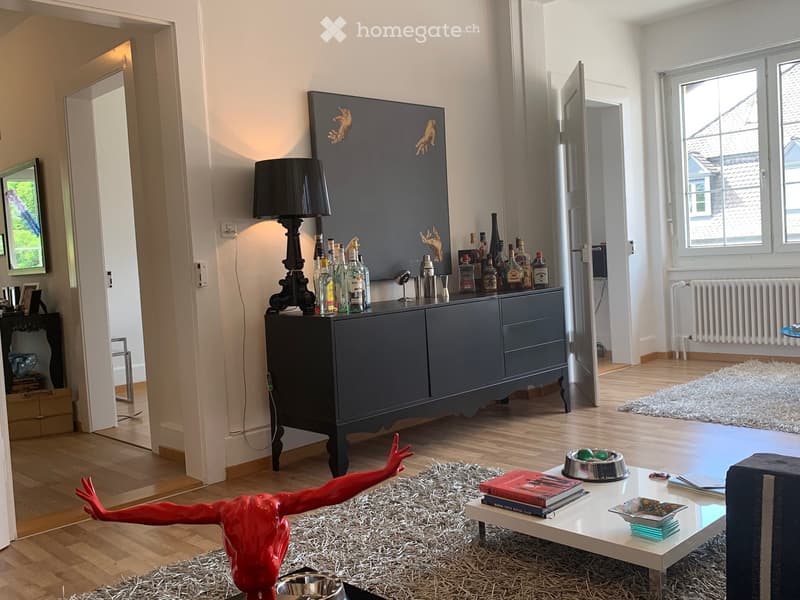 Modern living room with wooden flooring, a large dark grey cabinet, a floor lamp, a red sculpture, a coffee table with books and a bowl, and a mirror on the wall.