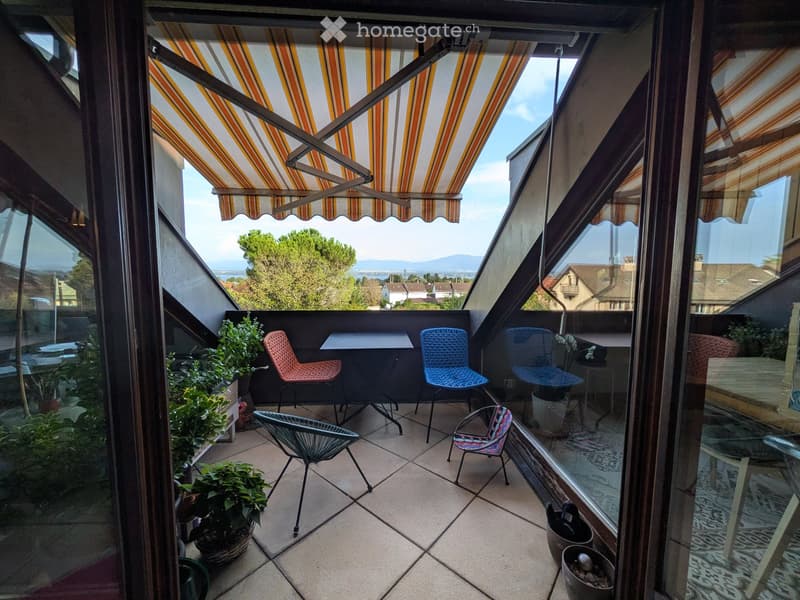 Balcony with a retractable awning, tiled floor, and several chairs and a table arranged for seating. Potted plants are placed around the area, and the view includes trees and a distant body of water.
