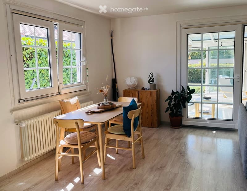 Bright dining area with wooden table, chairs, sideboard, vase with flowers, and potted plant near large windows and sliding glass door leading to outdoor space.