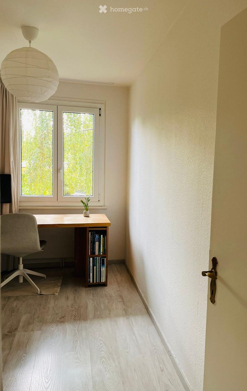 A small, well-lit room with a wooden floor, a desk with a chair, a bookshelf, and a small plant on the windowsill.