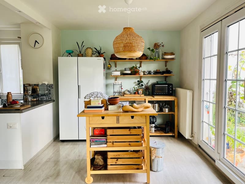 Bright kitchen with light green walls, white cabinets, and a wooden kitchen island. Features a pendant light, shelves with plants and dishes, and a refrigerator.