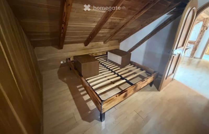An attic bedroom with wooden slanted ceilings, a wooden bed frame, and a tiled floor. The room is empty except for the bed frame and two boxes placed on it.