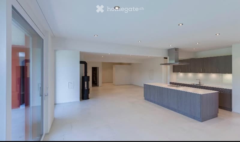 Modern, minimalist kitchen and living area with white walls, light wood flooring, and a large island. The space is completely unfurnished, showing only the structural elements and built-in cabinetry.