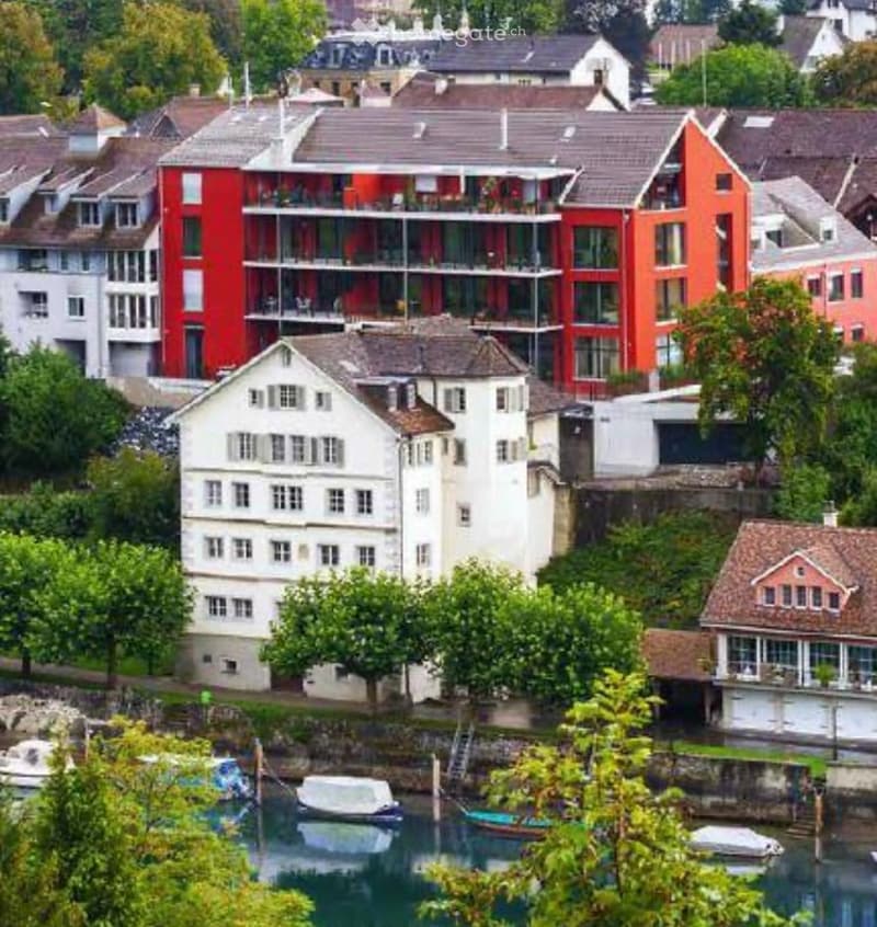 A multi-story residential building with red and white facades, surrounded by other buildings and greenery, with a waterfront featuring docked boats.