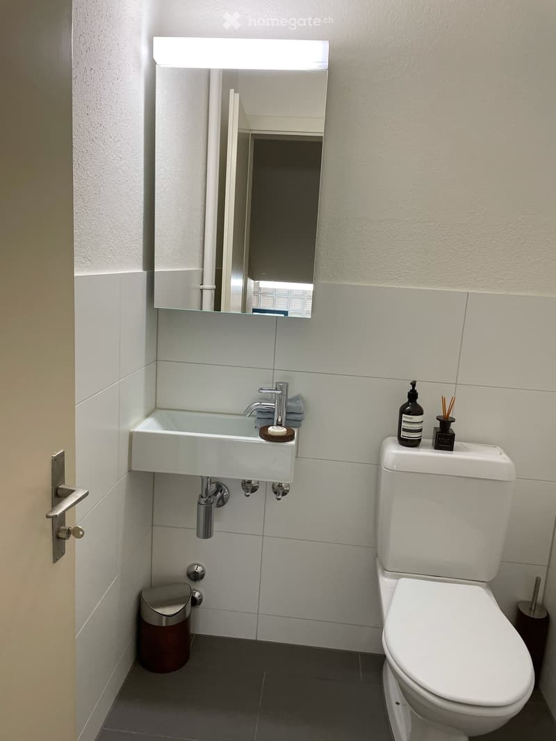 Modern bathroom with white tiles, a sink with a chrome faucet, a toilet, a mirror with built-in lighting, and a trash can.