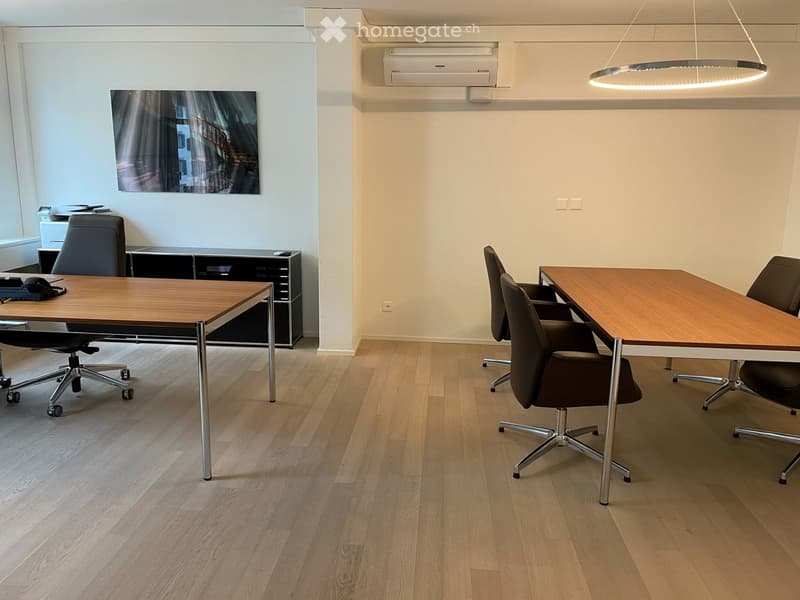 Modern office space with light wood flooring, two office tables, several chairs, a cabinet, and a large circular light fixture.