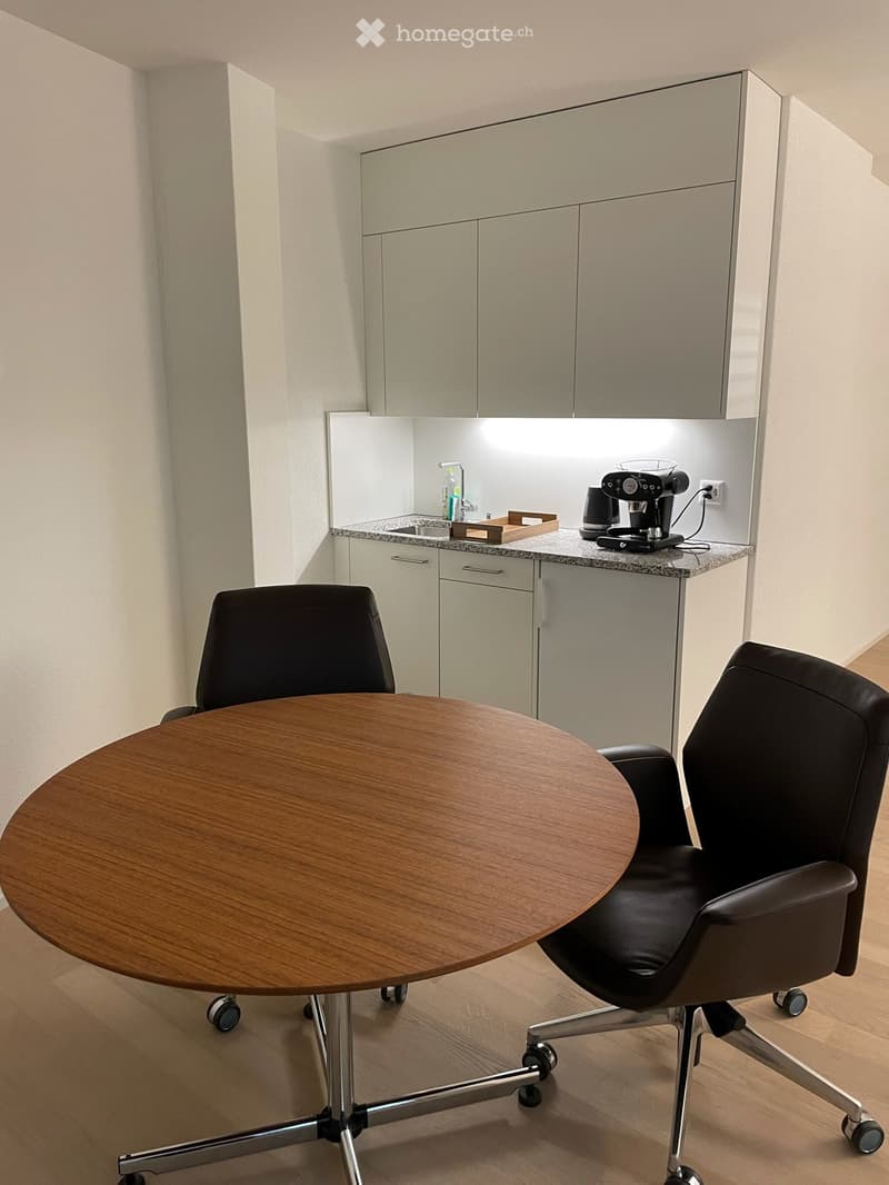 A modern kitchen area with a round wooden table, two black office chairs, a coffee machine, a sink with a faucet, and light-colored cabinets.