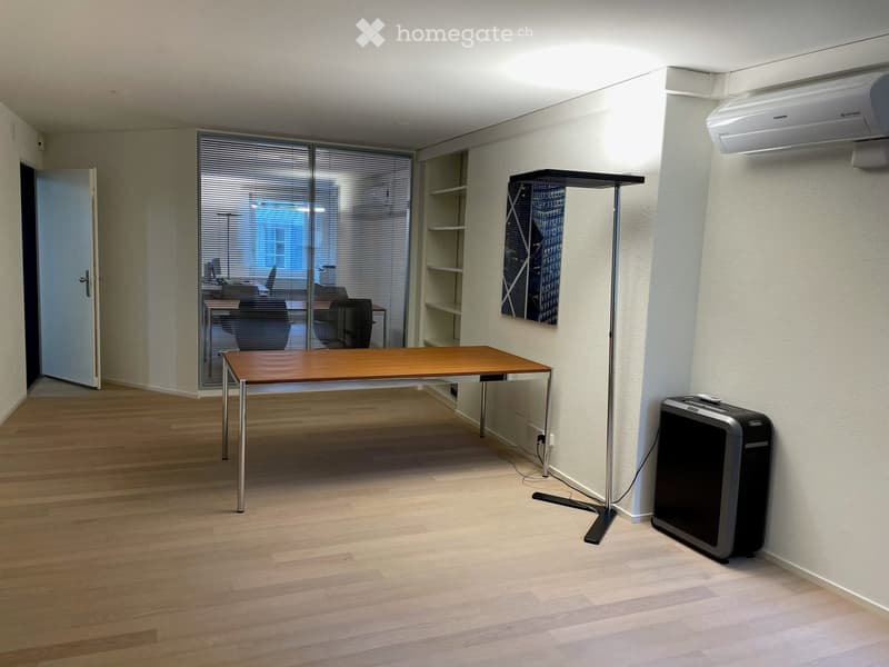 A modern, minimalist room with a wooden floor, a large wooden table, a chair, a floor lamp, a shelf, an air conditioner, and a fan.