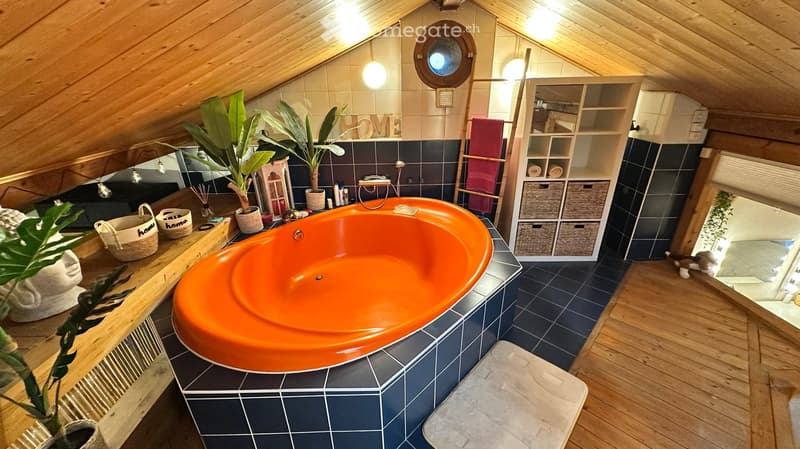Cozy bathroom with a large orange bathtub, dark blue tiles, wooden ceiling beams, and a round window. Includes a shelf with towels, a plant, and decorative items.