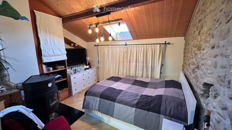 Cozy attic bedroom with wooden slanted ceilings, skylight, stone wall, bed with checkered quilt, TV, dresser, bookshelf, and fireplace.