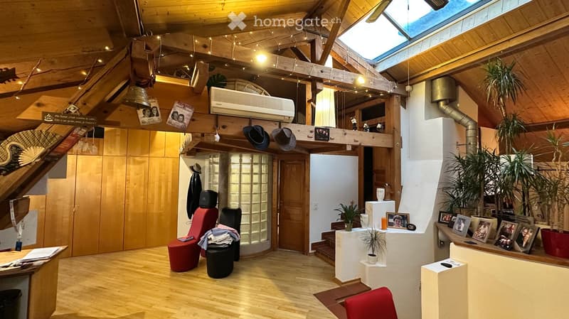 A spacious, well-lit room with wooden beams and a skylight, featuring a red chair, a black stool, a small table with plants, and several picture frames on a counter.
