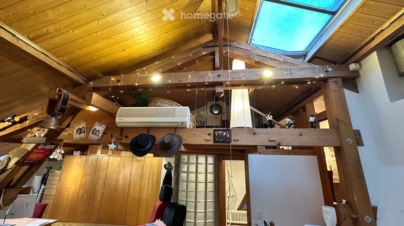 A cozy, wooden-framed room with exposed beams, a skylight, and various decorative items including hats, pictures, and a guitar. There is a table, chair, and air conditioner visible.