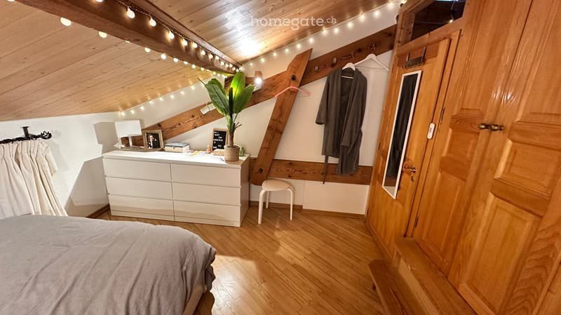 Cozy attic bedroom with wooden slanted ceilings, exposed beams, and warm lighting. Features a bed, white dresser, small stool, mirror, and potted plant.