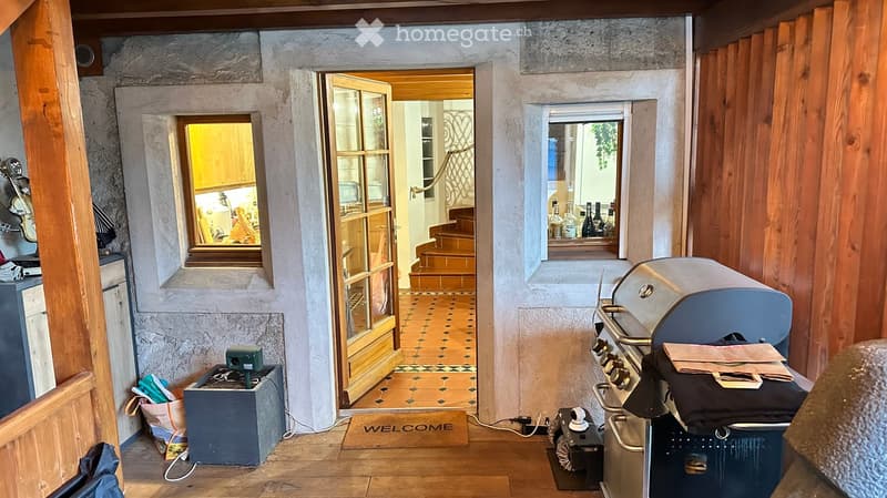 The image shows a cozy, rustic hallway with wooden floors and exposed stone walls. A grill is positioned on the right, with a guitar leaning against the wall nearby. A welcome mat lies on the floor, and a small table with a camera and other items is visib