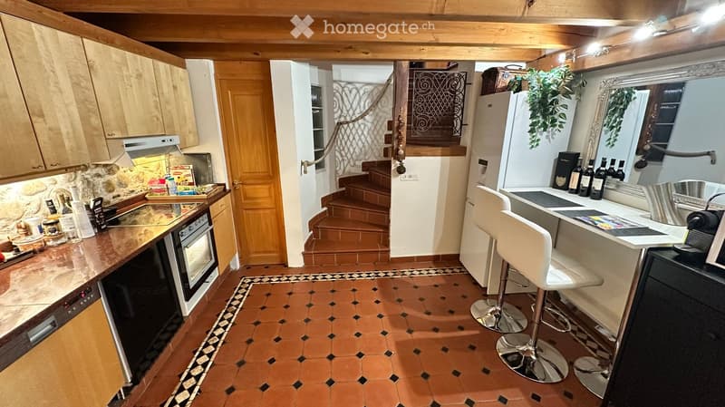 The image shows a cozy kitchen with wooden cabinets, a stone backsplash, and a tiled floor. There is an oven, refrigerator, and a small bar area with stools. The kitchen also features a staircase leading to another level.