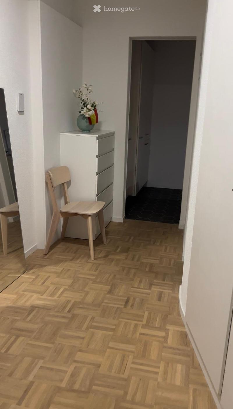 A narrow hallway with wooden herringbone flooring, white walls, and a small side table with a vase of flowers and a flag. A simple wooden chair is placed near the table, and a doorway leads to another room.