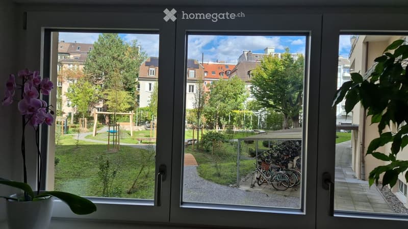 View from a balcony showing a green park area with trees, playground equipment, and a paved path with bicycles parked nearby.